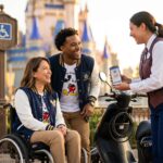 Wheelchair user and companion at Walt Disney World entrance with Cast Member showing a DAS return time on a smartphone, ECV parked nearby, castle landmark in background